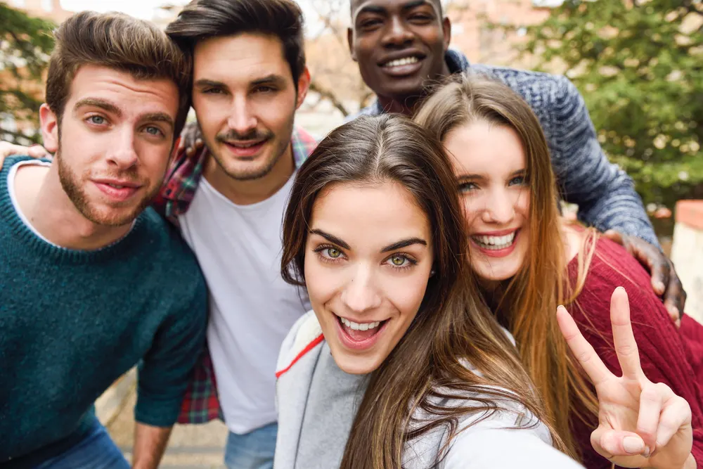 A group of five friends smiling and posing for a selfie outdoors. The person in front is making a peace sign with their fingers. Trees and the blurred background of a street can be seen behind them. Everyone appears happy and close together. A group of five friends smiling and posing for a selfie outdoors. The person in front is making a peace sign with their fingers. Trees and the blurred background of a street can be seen behind them. Everyone appears happy and close together.
