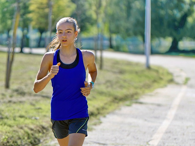 A young person with long hair in a ponytail, wearing a blue tank top and black shorts, runs on a paved path in a park. The background is green with trees, and the weather appears to be sunny. A young person with long hair in a ponytail, wearing a blue tank top and black shorts, runs on a paved path in a park. The background is green with trees, and the weather appears to be sunny.